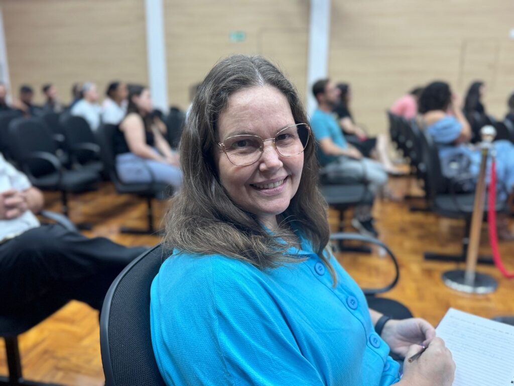 A mulher na foto está sorrindo e olhando para a câmera. Ela tem cabelos castanhos longos e usa óculos de aros dourados. Ela está vestindo uma blusa azul brilhante com botões. Ela está sentada em uma cadeira preta, e em suas mãos ela segura um caderno e uma caneta, como se estivesse fazendo anotações. Ao fundo, há um grupo de pessoas sentadas em cadeiras, parecendo estar em uma palestra ou evento. O ambiente parece ser uma sala de conferências ou auditório, com paredes de madeira e piso de madeira clara. A iluminação é suave, criando uma atmosfera acolhedora.