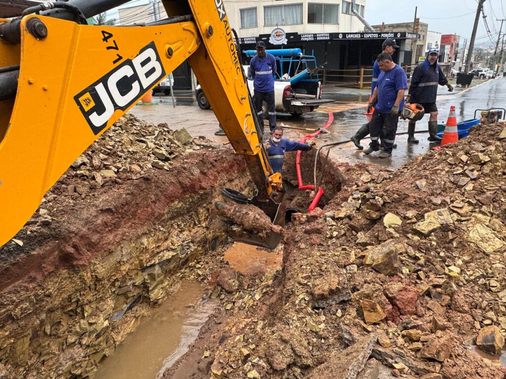 A imagem mostra uma cena de trabalho em uma rua, com uma escavadeira JCB amarela em primeiro plano. A escavadeira está em processo de escavação de uma vala profunda e lamacenta, onde se vêem tubulações de cor vermelha e um trabalhador dentro da vala, aparentemente supervisionando ou auxiliando na instalação. Ao redor da vala, há montes de terra e pedras removidas. Vários trabalhadores estão presentes na cena, alguns vestindo uniformes azuis e outros com roupas de trabalho mais casuais. Um dos trabalhadores carrega uma motosserra. Ao fundo, observa-se uma rua molhada, possivelmente devido à chuva, com edifícios comerciais e outros veículos. Há também cones de trânsito laranja indicando a área de trabalho. A atmosfera geral sugere uma obra pública ou de infraestrutura em andamento.