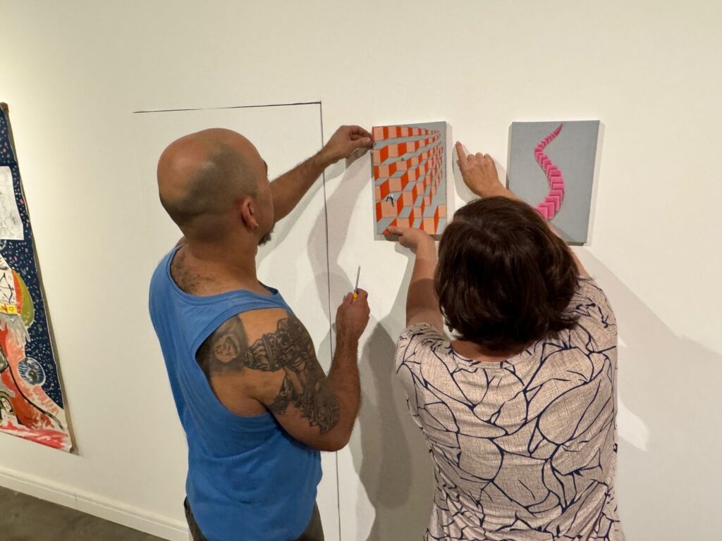 A imagem mostra duas pessoas pendurando quadros em uma parede branca. À esquerda, um homem careca com uma camiseta azul sem mangas e tatuagens no braço esquerdo está ajustando um quadro com uma chave de fenda na mão. O quadro que ele está ajustando tem um design abstrato com quadrados laranja e cinza, e uma pequena figura humana no centro. À direita, uma mulher com cabelo castanho escuro e uma blusa bege com um padrão de linhas escuras está apontando para outro quadro. Este segundo quadro, à direita do primeiro, tem um fundo cinza e uma forma abstrata rosa que se assemelha a uma serpente ou corrente. Ao lado esquerdo do homem, vislumbra-se parte de um quadro maior, com um design mais elaborado e colorido, com elementos que lembram um mapa astral ou um diagrama complexo. A iluminação na cena sugere um ambiente de galeria de arte ou exposição.
