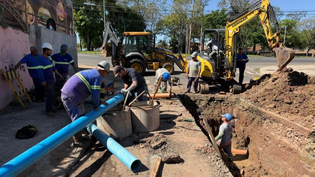 A imagem mostra uma equipe de trabalhadores em um canteiro de obras, instalando tubulações azuis em uma vala. Há duas escavadeiras no local, uma maior amarela da marca DEERE e uma menor amarela da marca YANMAR. Vários trabalhadores, vestidos com uniformes azuis e coletes de alta visibilidade, estão engajados em diferentes tarefas, como manusear os tubos, operar equipamentos e supervisionar o trabalho. O ambiente sugere um trabalho de infraestrutura, possivelmente de saneamento ou abastecimento de água, em uma área urbana, com uma rua e construções ao fundo.