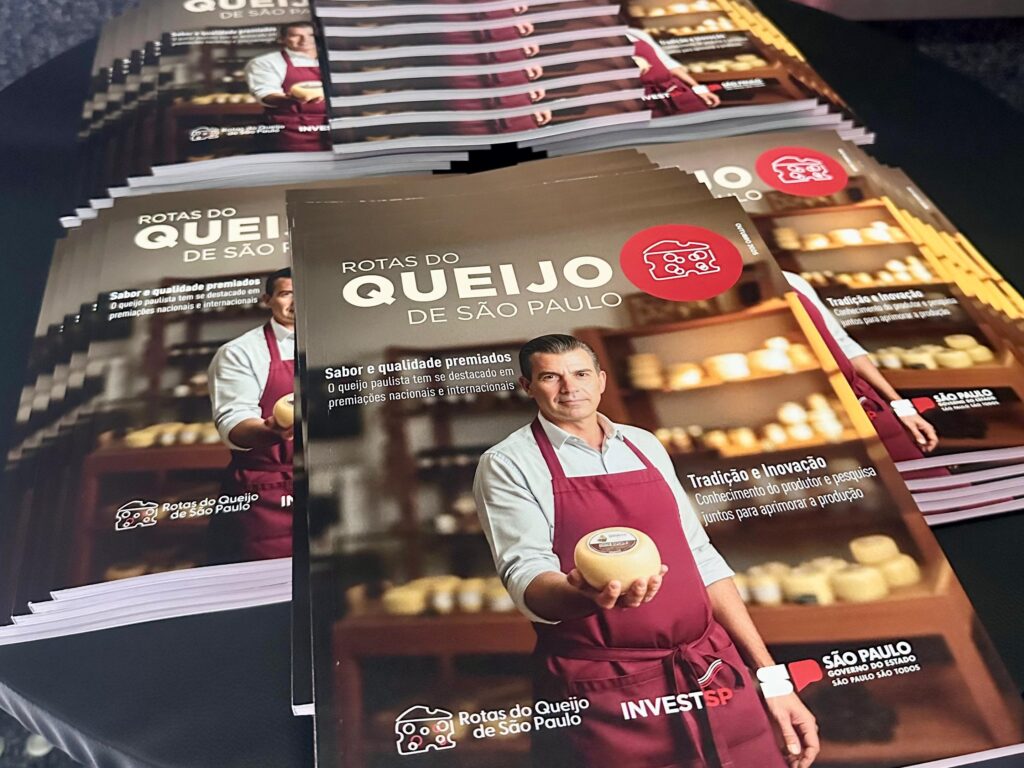 A imagem mostra uma pilha de folhetos sobre o tema "Rotas do Queijo de São Paulo". Os folhetos estão dispostos de forma escalonada, com o folheto da frente em destaque. Ele exibe a imagem de um homem sorridente, vestindo um avental vermelho e segurando um queijo redondo. O título "Rotas do Queijo de São Paulo" está em letras grandes e proeminentes. Abaixo do título, há uma descrição sobre "Sabor e qualidade premiados", indicando que o queijo paulista tem se destacado em premiações nacionais e internacionais. Há também um ícone de queijo vermelho e o logo "INVESTSP". Outros folhetos na pilha também mostram o mesmo conteúdo, alguns ligeiramente visíveis nas bordas. A iluminação da foto sugere um ambiente interno, possivelmente uma feira ou evento relacionado ao setor de laticínios.