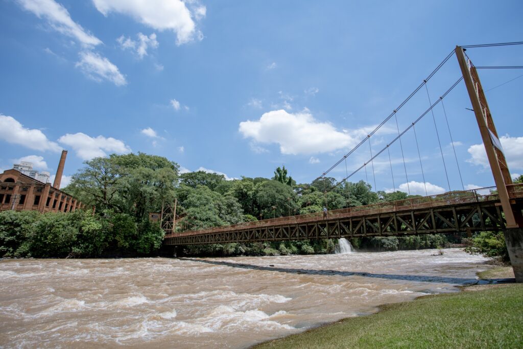 A imagem retrata uma ponte suspensa antiga, com estrutura de metal enferrujado, cruzando um rio de águas barrentas e agitadas. Ao fundo, uma densa floresta verde e, à esquerda, um edifício de tijolos vermelhos com uma chaminé alta, possivelmente uma antiga fábrica. O céu azul, com algumas nuvens brancas e fofas, completa a paisagem, transmitindo uma sensação de nostalgia e imponência. A ponte parece ser um ponto de conexão entre o passado industrial e a natureza exuberante.