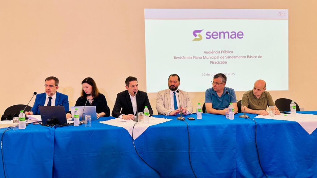 A imagem mostra uma audiência pública realizada para a revisão do Plano Municipal de Saneamento Básico de Piracicaba, datada de 16 de outubro de 2025. Seis pessoas estão sentadas lado a lado em uma mesa coberta por um pano branco com a logo "semae" em azul. Atrás deles, há um telão projetando o tema do evento. As pessoas parecem ser autoridades ou especialistas, vestidas de forma formal, com microfones à frente, sugerindo que estão participando de uma discussão ou apresentação. Há garrafas de água e copos sobre a mesa. O ambiente aparenta ser uma sala de conferências ou auditório, configurado para um evento oficial ou institucional.