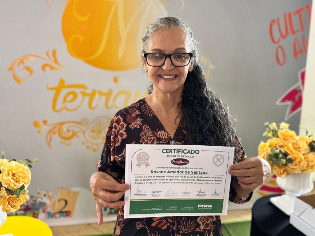 A imagem mostra uma mulher sorridente, com cabelos grisalhos e óculos, segurando um certificado. O certificado é referente a um curso de pizzaiolo, realizado pela Prefeitura de Piracicaba, e está emoldurado por um fundo que parece ser uma parede decorada com algumas palavras e logotipos, incluindo "terravia" e "CULT O A". O certificado confirma que a aluna Rosane Amador de Santana concluiu o curso entre 11 de setembro e 9 de outubro de 2025, com carga horária de 16 horas. A imagem transmite um momento de conquista e celebração pelo término do curso.