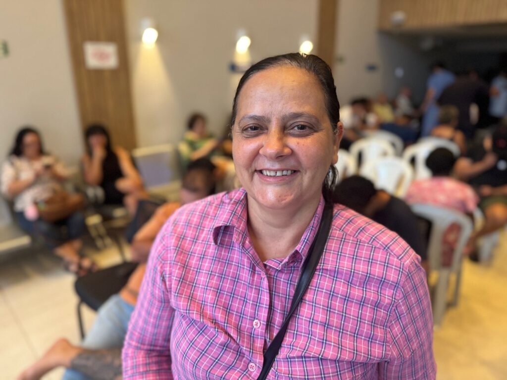 A mulher sorri para a câmera, vestindo uma camisa xadrez rosa e roxa. Ela tem cabelos escuros presos para trás e usa uma bolsa preta a tiracolo. O fundo está desfocado, mas parece ser uma sala com várias pessoas sentadas em cadeiras, possivelmente em uma sala de espera ou evento. Há luzes de teto e uma parede de madeira no fundo.