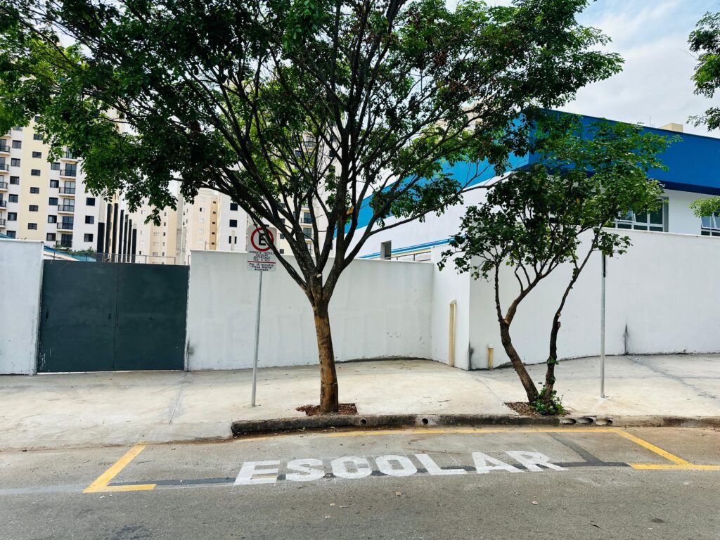 A imagem mostra uma rua com a palavra "ESCOLAR" pintada em letras brancas grandes no asfalto. Há uma calçada com duas árvores, uma maior e outra menor, e uma parede branca ao fundo. Atrás da parede branca, vê-se um edifício alto com muitas janelas e um prédio azul com janelas brancas. Um sinal de trânsito com um "E" vermelho dentro de um círculo está próximo à árvore maior. Há linhas amarelas pintadas na rua e na calçada.