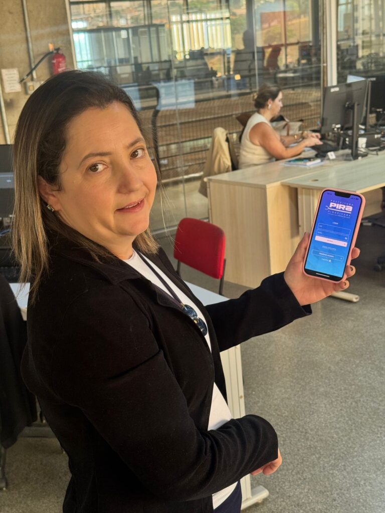 A imagem mostra uma mulher sorrindo para a câmera, segurando um celular. O celular exibe um aplicativo com o logotipo "PREFEITURA PIRACICABA" e campos para login. A mulher está em um ambiente de escritório, com outras pessoas e computadores ao fundo. Ela veste uma jaqueta preta sobre uma blusa branca.