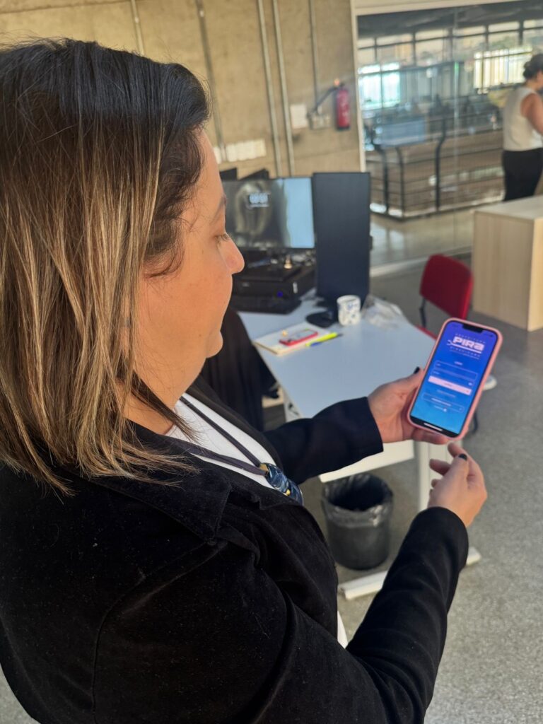 A mulher, vista de perfil, segura um smartphone rosa em sua mão direita. A tela do telefone exibe um aplicativo com o logotipo "PIRA" na parte superior e campos para login. Ela está usando uma jaqueta preta sobre uma blusa branca e tem cabelos escuros com mechas loiras. Ao fundo, um escritório com mesas, computadores e uma porta de vidro que dá para outro ambiente.