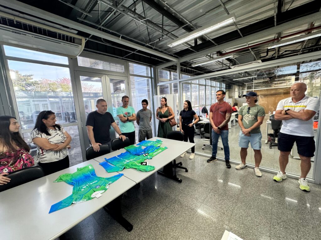 A imagem mostra um grupo de pessoas em uma sala de reuniões, com várias camisetas dobradas sobre uma mesa. As camisetas são predominantemente verdes e azuis, com um design tie-dye. Há oito pessoas visíveis na sala, em pé e sentadas. As janelas grandes na lateral da sala deixam entrar luz natural e mostram a vegetação do lado de fora. O ambiente parece ser um escritório ou espaço de trabalho moderno, com piso de concreto polido e teto industrial. As pessoas estão vestidas de forma casual, sugerindo um ambiente descontraído. Algumas pessoas estão olhando para as camisetas na mesa, enquanto outras conversam entre si.