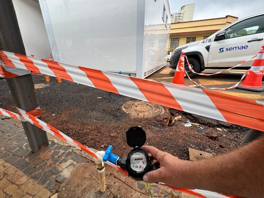 A imagem mostra uma instalação de um hidrômetro em uma área externa, possivelmente em uma obra ou manutenção. * Um hidrômetro preto e branco, com a tampa aberta, está sendo segurado por uma mão. O mostrador do hidrômetro exibe o número 000005 m³. Uma válvula azul está conectada ao hidrômetro. * Uma fita de isolamento laranja e branca está estendida na frente da cena, separando a área de trabalho. * Ao fundo, há um módulo branco modular, cones de trânsito laranja e um veículo branco com o logotipo "semae" e o número 478. * O chão ao redor é composto por terra solta e pedras, com algumas tampas de bueiro visíveis. * Uma calçada de pedras irregulares está presente no lado esquerdo da imagem. * O céu está nublado, indicando um dia com pouca luz solar.