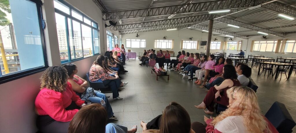 A imagem mostra um grupo de pessoas, predominantemente mulheres, reunidas em um espaço interno que parece ser um refeitório ou auditório. Elas estão sentadas em cadeiras dispostas em um círculo, com algumas pessoas em pé na frente, possivelmente apresentando ou conduzindo uma reunião. Muitas das pessoas na imagem estão vestindo roupas na cor rosa, sugerindo que o evento pode estar relacionado a alguma campanha ou celebração, como o Outubro Rosa. O ambiente possui janelas grandes que permitem a entrada de luz natural, e o teto é metálico com vigas aparentes e luminárias. Ao fundo, há mesas e cadeiras de refeitório, indicando o uso multifuncional do espaço. A atmosfera parece ser de atenção e participação, com a maioria das pessoas voltadas para o centro do círculo.