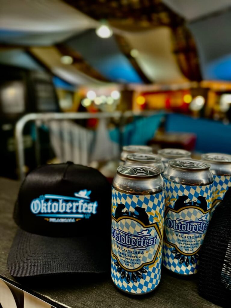 A imagem mostra uma mesa com várias latas de cerveja e um boné, todos com a temática do Oktoberfest. As latas de cerveja são prateadas e apresentam um design xadrez azul e branco, com o logo "Oktoberfest" em destaque. Abaixo do logo, lê-se "PIRACICABA", "CERVEJA ESTILO OKTOBERFEST", o teor alcoólico de 5,3% e o volume de 473 ml. Ao lado das latas, há um boné preto com o mesmo logo "Oktoberfest" bordado em azul e branco. O boné está sobre outro boné, também preto, que não é totalmente visível. O fundo da imagem está desfocado, mas sugere um ambiente festivo com tecidos coloridos pendurados no teto e luzes.