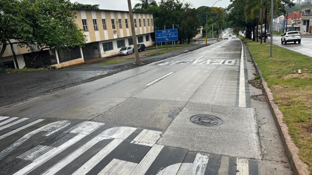A imagem mostra uma rua em um dia nublado, com o asfalto molhado, possivelmente devido à chuva. Há uma faixa de pedestres em primeiro plano, com as marcações brancas desgastadas em algumas partes. A rua é larga e parece ser uma via importante, com várias pistas de rolamento. À esquerda da rua, há um edifício de dois andares com janelas retangulares, construído em alvenaria aparente com detalhes em branco. Uma árvore frondosa com galhos que se estendem sobre a calçada está próxima ao edifício. Uma cerca escura com barras diagonais separa a propriedade da rua. À direita da rua, há um gramado bem cuidado que desce em direção à via. Mais adiante, percebe-se a presença de outras casas e edifícios, com um estilo arquitetônico mais residencial. Vários carros estão circulando na rua, e um deles, um SUV branco, está em destaque mais ao fundo, movendo-se na direção oposta ao observador.