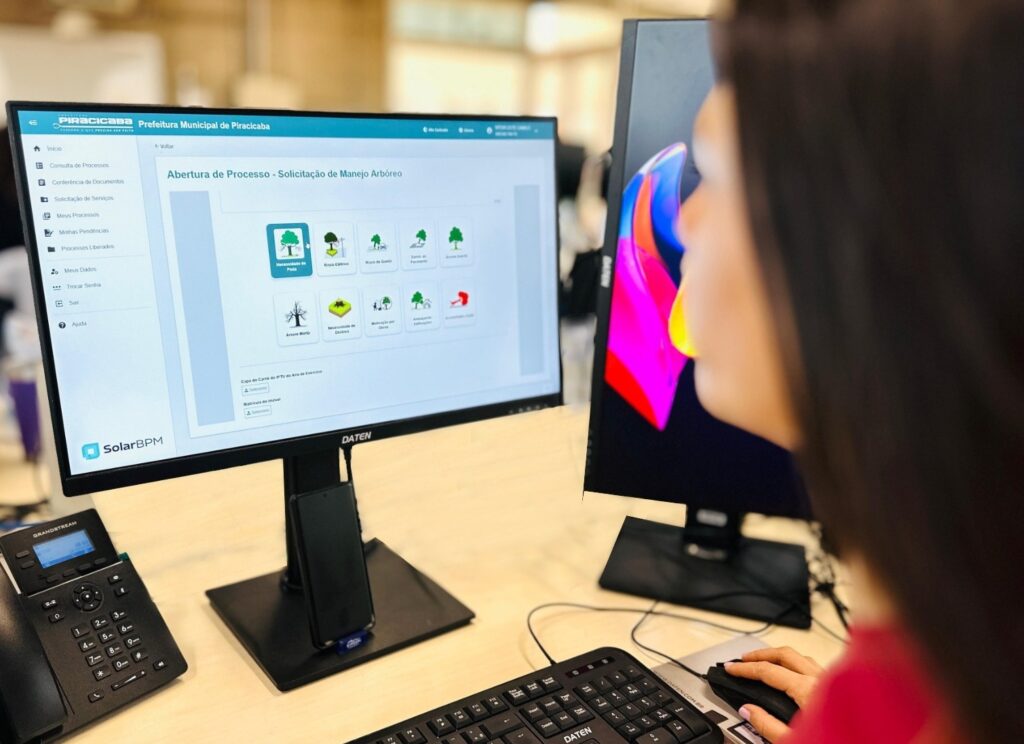 A imagem mostra uma mulher trabalhando em um computador em um escritório. Ela está usando um sistema online da Prefeitura Municipal de Piracicaba para solicitar o manejo arbóreo. A tela do computador exibe um formulário com diversas opções para o tipo de solicitação, como "Necessidade de Poda", "Risco Elétrico", "Risco de Queda", entre outras. Há também campos para preencher informações como "Capa do Carnê do IPTU do Ano de Exercício" e "Matrícula do Imóvel". Ao lado do monitor principal, há outro monitor com uma imagem colorida e abstrata. Na mesa, também é possível ver um telefone IP da marca Grandstream, um teclado e um mouse. A mulher está focada na tela, com a mão no mouse, indicando que está ativamente utilizando o sistema.