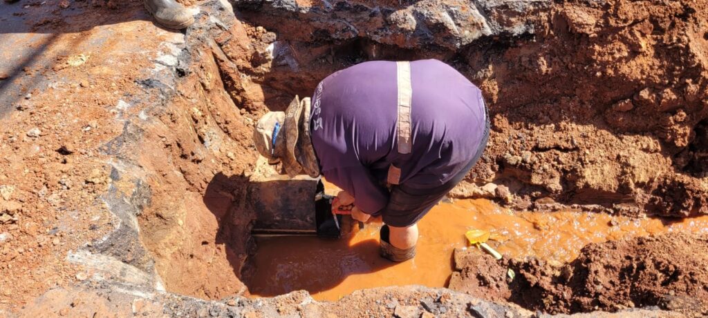 A imagem mostra um trabalhador agachado em uma vala cavada no solo, que parece ser de terra avermelhada e pedras. O trabalhador está vestindo uma camisa roxa com uma faixa refletiva, calças escuras e botas de borracha. Ele está focado em uma tubulação escura que está parcialmente submersa em água lamacenta e de cor alaranjada. Ao lado do trabalhador, na água, pode-se ver uma pequena pá amarela. As paredes da vala são irregulares e mostram camadas de terra e, possivelmente, asfalto ou algum outro material escuro na parte superior. A iluminação sugere que a foto foi tirada durante o dia, com luz solar direta. A cena indica um trabalho de reparo ou manutenção em uma infraestrutura subterrânea.