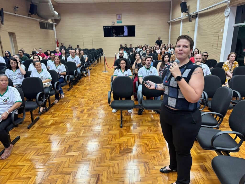 A imagem mostra uma sala de conferências com um público sentado em cadeiras, voltados para a frente, onde uma mulher está de pé, segurando um microfone e um controle remoto. Ela está de frente para a audiência, parecendo estar no meio de uma apresentação ou discurso. A sala tem um piso de madeira com um padrão de espinha de peixe e paredes de madeira clara. Há um projetor e uma tela na frente da sala, e um retrato pendurado acima da tela. A audiência é composta por homens e mulheres de várias idades, a maioria vestindo camisetas brancas com um logotipo verde. A mulher que está de pé usa um colete xadrez sobre uma blusa preta e calças pretas. A iluminação na sala é suave e uniforme.