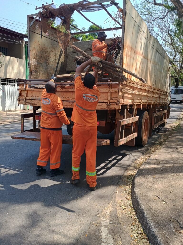 Na imagem aparecem três homens com uniforme laranjado, carregando um caminhão com restos de madeira. Dois homens estão embaixo no chão e o outro de boné recolhe os objetos. O caminhão está estacionado em uma rua e sobre ele aparece um árvore com poucos galhos.