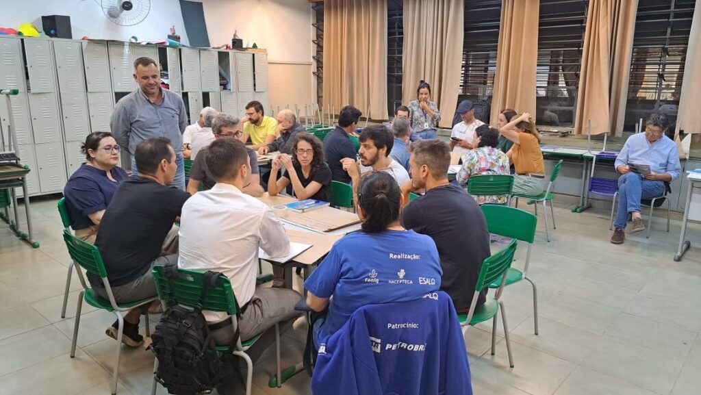 A imagem mostra um grupo de pessoas reunidas em uma sala, possivelmente para uma reunião ou workshop. Há várias mesas dispostas em círculo, e as pessoas estão sentadas ao redor delas, algumas conversando e outras olhando para papéis ou dispositivos. Ao fundo, há fileiras de armários metálicos, semelhantes aos encontrados em escolas ou vestiários. Há também cortinas nas janelas e um ventilador de teto. No primeiro plano, vemos algumas pessoas sentadas em cadeiras verdes. Uma mulher com óculos está sentada à esquerda, vestindo uma blusa azul escura. Ao lado dela, um homem de camisa preta olha para frente. Outras pessoas estão sentadas em frente a eles, com suas costas voltadas para a câmera. Uma delas usa uma camiseta azul com um texto escrito em branco, que parece ser "Realização: FENIQ NACEPTECA ESALQ". Abaixo, há outro texto que parece ser "Patrocínio: PETROBRAS".
