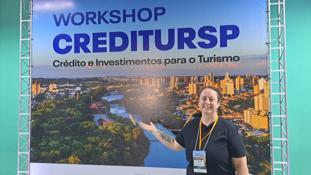 A imagem mostra uma mulher sorrindo em frente a um grande banner que anuncia o "WORKSHOP CREDITURSP". O banner exibe uma paisagem urbana com um rio e edifícios altos, com o texto em azul e branco. A mulher usa uma camiseta preta e um crachá pendurado no pescoço. Ela está com o braço estendido, apontando para o banner, e tem uma tatuagem visível no braço esquerdo. O fundo da imagem é um tom de verde claro.