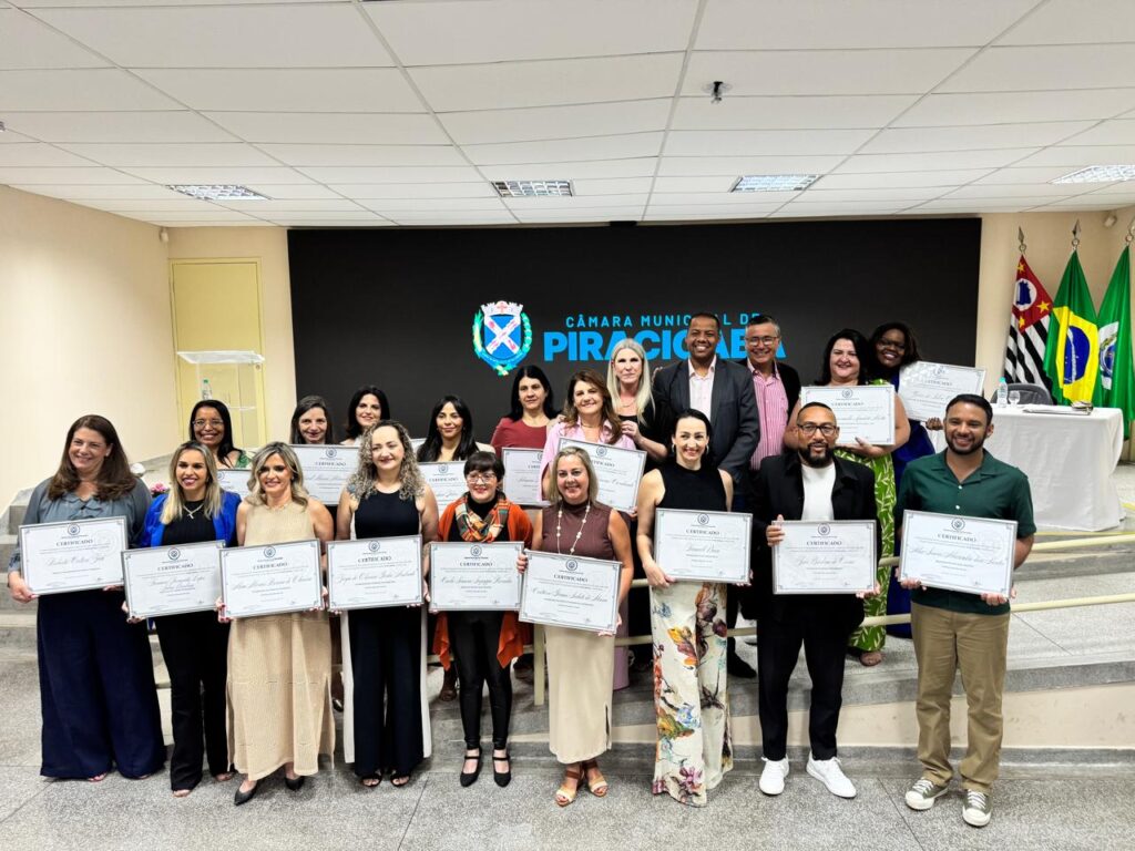 Um grupo de pessoas está reunido em um ambiente interno, participando de uma cerimônia formal na Câmara Municipal de Piracicaba. Vários deles seguram certificados, indicando que foram reconhecidos ou homenageados. Ao fundo, há um palco com um pódio, uma mesa coberta por um tecido verde e bandeiras do Brasil e do estado de São Paulo, reforçando o caráter oficial do evento. Todos estão vestidos com roupas sociais, e o clima é de celebração e reconhecimento público.