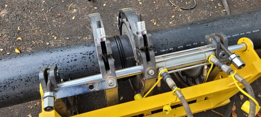 A imagem mostra um close-up de um equipamento de fusão de tubos de plástico, provavelmente para sistemas de água ou gás. Um tubo preto de grande diâmetro está posicionado horizontalmente, e o maquinário de fusão está montado sobre ele. O maquinário em si é composto por partes metálicas robustas, algumas pintadas de amarelo vibrante, que abraçam o tubo. Vemos elementos hidráulicos, como mangueiras pretas com conexões rápidas e um cilindro prateado, que provavelmente aplicam a pressão e o calor necessários para fundir as extremidades dos tubos. Há também peças de metal cinza com parafusos e molas visíveis, que parecem ser os mecanismos de fixação e ajuste.