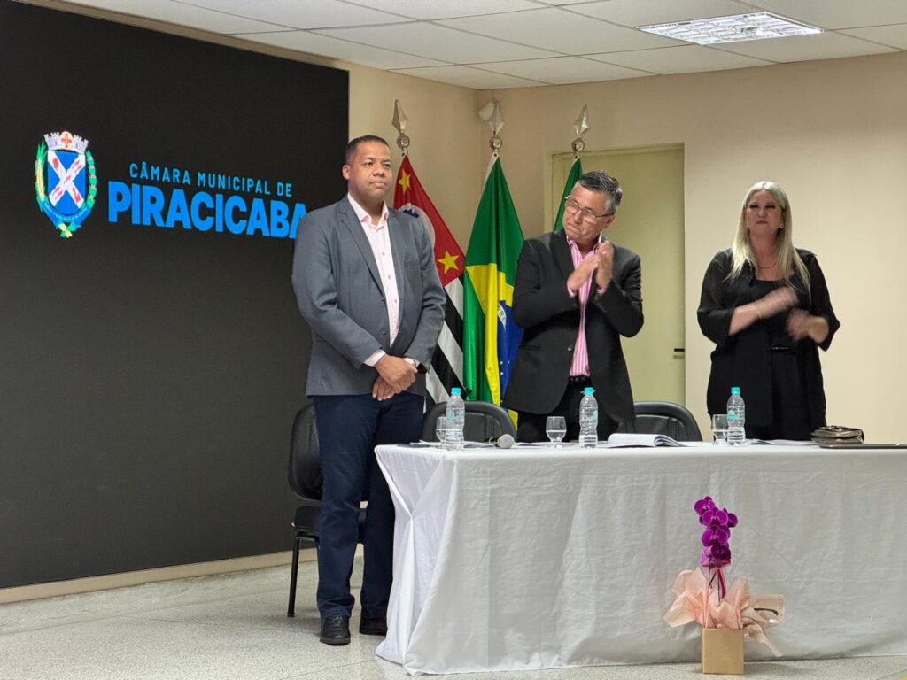 Pra cego ver: Três pessoas estão em pé atrás de uma mesa em um ambiente formal dentro da Câmara Municipal de Piracicaba. À esquerda, um homem está com as mãos cruzadas à frente do corpo; ao centro, outro homem aplaude; e à direita, uma mulher aplaude. Sobre a mesa há garrafas de água, microfone, documentos e um arranjo de flores. Atrás deles, há bandeiras do Brasil, do estado de São Paulo e uma terceira parcialmente visível.