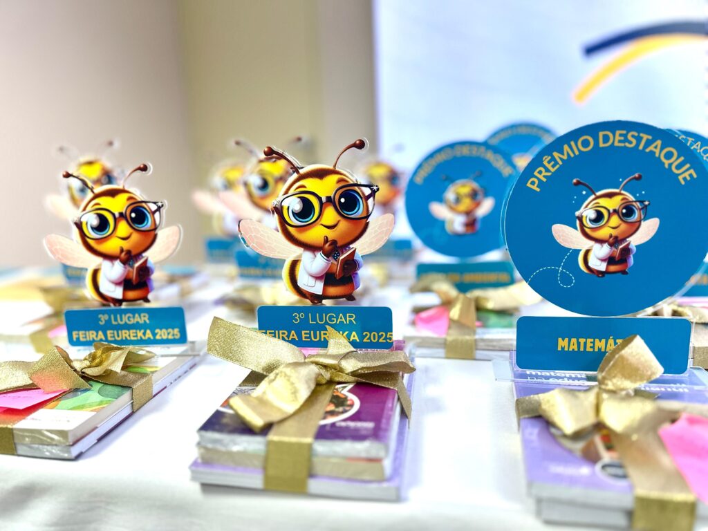 A imagem exibe uma coleção de prêmios com tema de abelha, organizados em uma mesa. As abelhas são personagens animados, usando óculos e jalecos de laboratório, segurando livros. Os prêmios consistem em pequenos livros ou cadernos amarrados com fitas douradas brilhantes. Cada prêmio tem um pequeno suporte azul com texto. Alguns suportes indicam "3º LUGAR FEIRA EUREKA 2025", enquanto outros exibem "PRÊMIO DESTAQUE" e "MATEMÁTICA". As abelhas em si estão em suportes transparentes, com algumas em forma de adesivos e outras em medalhões circulares azuis. O fundo é desfocado, mas sugere um ambiente interno, possivelmente uma sala de premiação ou exposição. A iluminação destaca os detalhes dourados das fitas e o brilho dos prêmios.