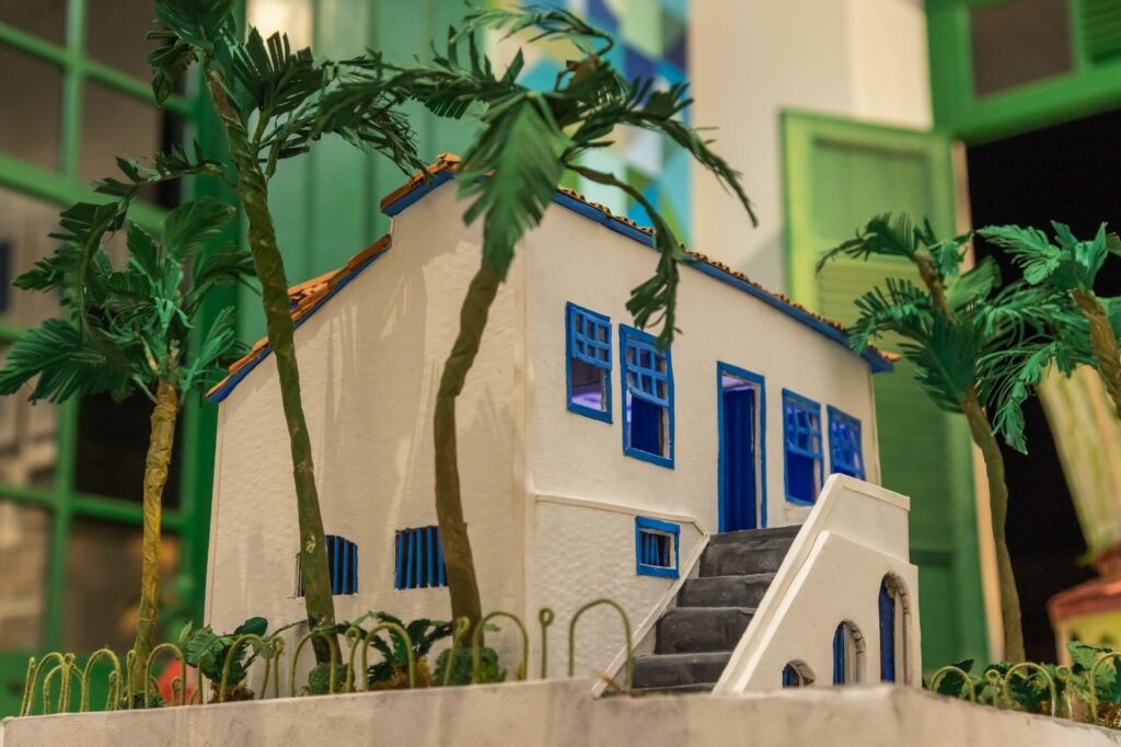 A imagem exibe uma miniatura detalhada de uma casa branca com telhado de telhas marrons e detalhes em azul vibrante. A casa possui uma escada que leva à porta principal e várias janelas, também em azul. Palmeiras com folhagem verde escura circundam a casa, adicionando um toque tropical à cena. O fundo sugere um ambiente externo com elementos verdes, possivelmente uma janela ou porta pintada de verde. As plantas na base da miniatura parecem ser feitas de papel ou material similar, com um estilo delicado e curvo. A iluminação suave realça os detalhes da maquete, criando uma atmosfera acolhedora e charmosa.