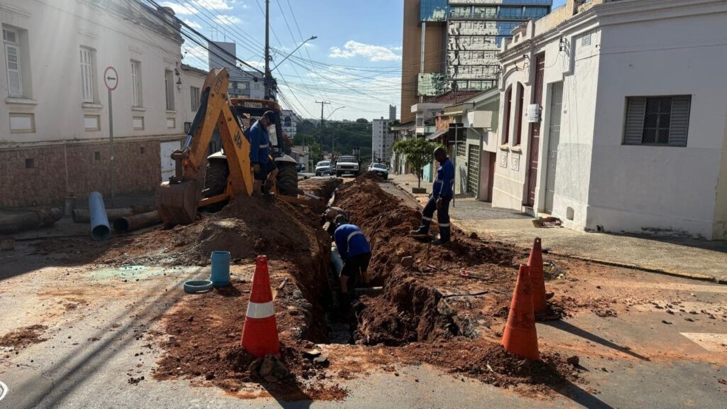 A imagem mostra uma cena de obras em uma rua urbana, com trabalhadores realizando reparos em tubulações subterrâneas. Um escavadeira amarela está posicionada ao lado de uma vala aberta na rua, com terra e detritos ao redor. Vários trabalhadores, vestindo uniformes azuis, estão engajados na tarefa, com alguns dentro da vala e outros supervisionando a operação. Tubos de diferentes tamanhos, incluindo alguns azuis e outros mais largos, estão empilhados nas proximidades, indicando o material que está sendo utilizado ou substituído. Cones de trânsito laranja e brancos foram colocados para sinalizar a área de trabalho e alertar os motoristas. Ao fundo, edifícios residenciais e comerciais compõem o cenário urbano, com destaque para um prédio moderno de vidro e um edifício mais antigo com detalhes arquitetônicos. A rua está parcialmente bloqueada pela obra, com alguns carros estacionados e em movimento ao longe. O céu está claro e azul, sugerindo um dia ensolarado. A imagem transmite a ideia de trabalho em andamento para a manutenção da infraestrutura urbana.