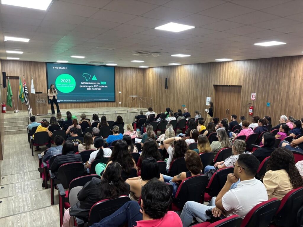 A imagem retrata uma sala de palestras com uma audiência sentada em cadeiras, voltada para um palco onde uma mulher está em pé, apresentando. Atrás dela, um grande telão exibe informações sobre Acidente Vascular Cerebral (AVC) no Brasil. O telão mostra o ano "2023" em destaque e a frase "Morrem mais de 300 pessoas por AVC todos os dias no Brasil", acompanhada de um mapa estilizado do Brasil. Há também um logotipo com as letras "SIMESPI" em um dos cantos do palco. A audiência é composta por pessoas de diversas idades, a maioria sentada em cadeiras dispostas em fileiras e organizadas em formato de auditório. A iluminação da sala parece ser artificial, vinda de luminárias no teto. Ao fundo, há paredes revestidas de madeira e algumas portas. Bandeiras nacionais e estaduais estão posicionadas à esquerda do palco.