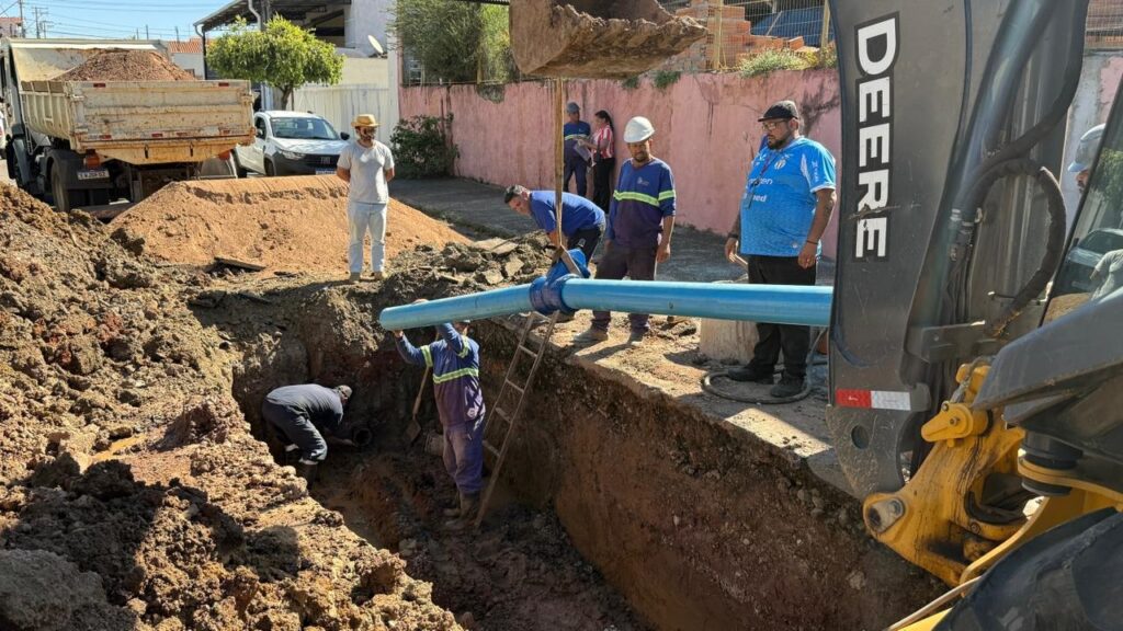 A imagem mostra uma equipe de trabalhadores em uma vala aberta, com o objetivo de instalar ou reparar uma tubulação de água azul. Há um caminhão basculante ao fundo, com uma carga de terra ou cascalho, e um trator John Deere ao lado da vala. Os trabalhadores estão vestidos com uniformes de trabalho, alguns com capacetes de segurança. Um deles está dentro da vala, trabalhando em uma conexão na tubulação, enquanto outros estão acima, ajudando a manobrar a tubulação e supervisionando o trabalho. A vala é profunda e a terra ao redor é visivelmente removida. A cena sugere um projeto de infraestrutura em andamento em uma área urbana, com casas e árvores visíveis ao fundo.
