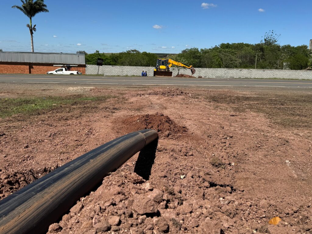 A imagem mostra uma cena de construção em um dia ensolarado. No primeiro plano, um grande tubo preto está parcialmente enterrado em solo vermelho e solto, com montes de terra ao redor. Ao fundo, uma estrada pavimentada separa a área de construção do outro lado. Do lado esquerdo da estrada, um prédio com uma parede de tijolos vermelhos e um telhado metálico cinza está visível, com uma palmeira alta ao lado. Um caminhão branco está estacionado perto do prédio. Mais adiante, à direita, um escavador amarelo está em operação, removendo terra. Duas pessoas estão sentadas perto da escavadeira. Uma parede de blocos de concreto cinza se estende ao longo da estrada, com árvores verdes e folhagens atrás dela. O céu está azul claro com algumas nuvens brancas dispersas.