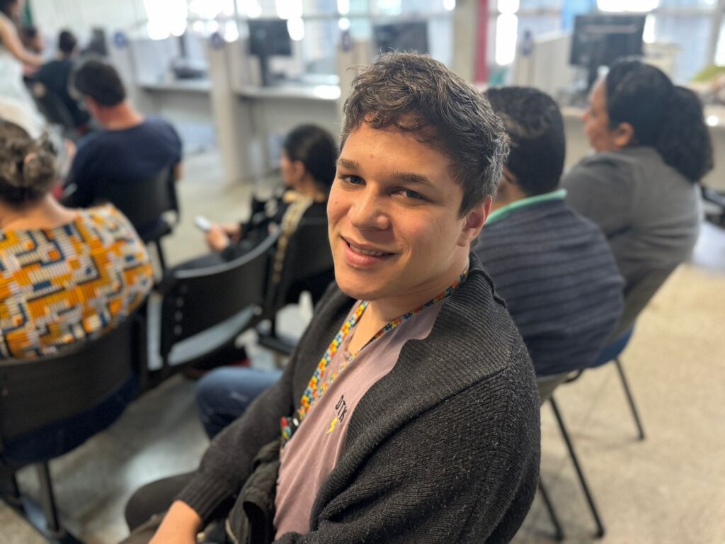 A imagem mostra um jovem sorrindo para a câmera, com outras pessoas sentadas em cadeiras desfocadas ao fundo. Ele está usando um suéter escuro sobre uma camiseta clara com um colar colorido. O ambiente parece ser um escritório ou sala de espera, com mesas e computadores visíveis ao fundo. A iluminação é suave e a composição foca no rosto do jovem.