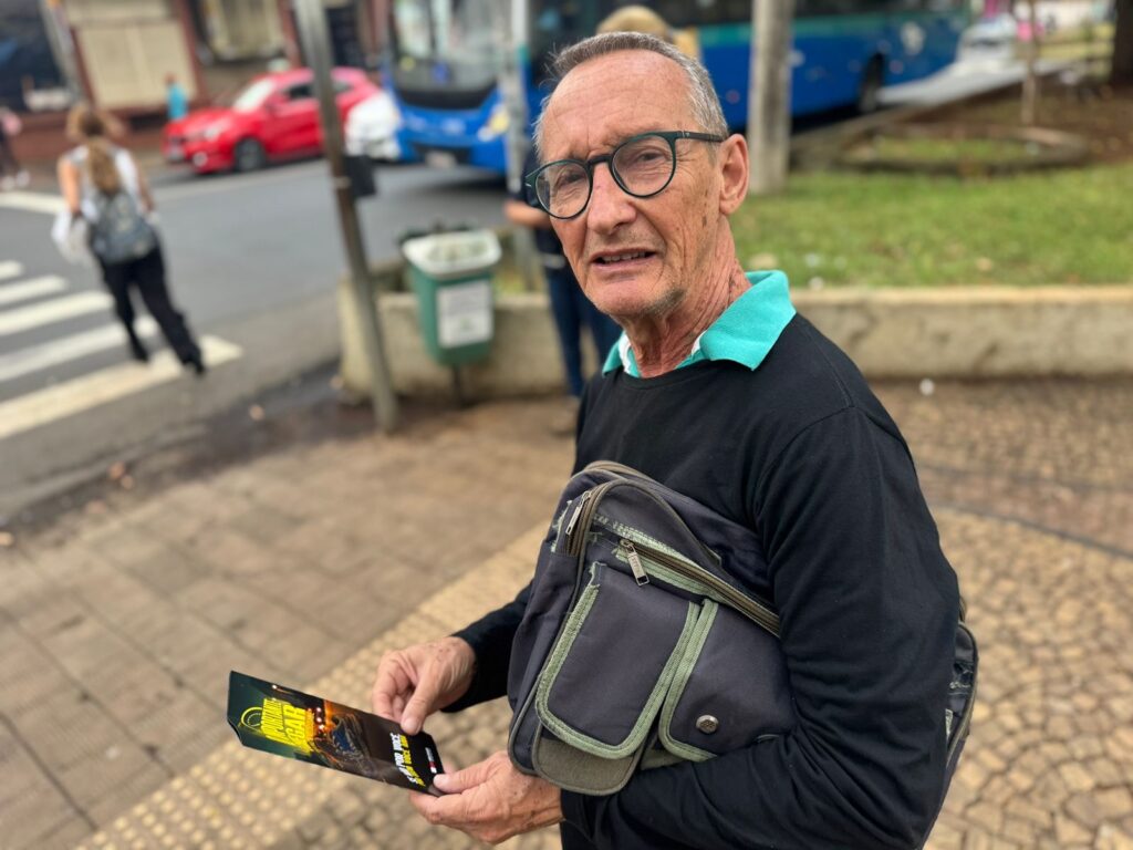 Um homem mais velho, com óculos de aro verde e cabelo grisalho, olha para a câmera. Ele está usando uma camiseta preta com gola verde por baixo. Ele está segurando um panfleto dobrado em suas mãos e tem uma bolsa escura a tiracolo. Ao fundo, vê-se uma rua movimentada com carros vermelhos e azuis, um ônibus e pessoas atravessando a rua em uma faixa de pedestres. Há também uma lixeira verde no lado direito da imagem. O homem parece estar em uma parada de ônibus ou em uma área pública onde as pessoas circulam. A iluminação sugere que é dia, possivelmente nublado.