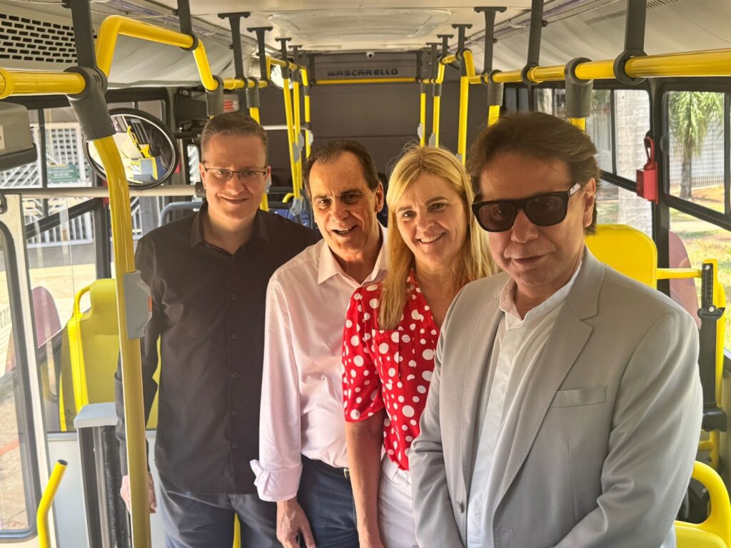 A imagem mostra quatro pessoas em pé dentro de um ônibus. Da esquerda para a direita, um homem com óculos escuros e camisa preta; um homem com camisa branca; uma mulher com uma blusa vermelha de bolinhas brancas; e um homem com óculos escuros e um blazer claro. Todos estão sorrindo e olhando para a câmera. O interior do ônibus é visível, com barras de apoio amarelas e assentos amarelos ao fundo. A iluminação sugere que a foto foi tirada durante o dia.