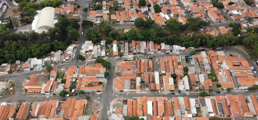 A imagem aérea mostra um bairro residencial com muitas casas de telhado vermelho. As casas são compactas e agrupadas, com ruas estreitas entre elas. Há muita vegetação, com árvores e arbustos em meio às casas e ao longo das ruas. Em destaque, uma grande estrutura com um telhado branco curvo, possivelmente um ginásio ou centro comunitário, está cercada por árvores. Vários carros estão estacionados em frente a essa estrutura. A paisagem urbana é densa, com uma mistura de casas de diferentes tamanhos e estilos, algumas parecendo mais antigas e outras mais novas. Algumas casas têm pátios ou áreas externas visíveis. A cena geral sugere uma área urbana consolidada, com um forte elemento natural presente em meio à construção.