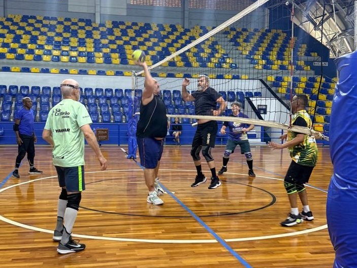 A imagem mostra um grupo de homens jogando vôlei em uma quadra interna. A quadra tem piso de madeira e arquibancadas azuis e amarelas ao fundo. No primeiro plano, um homem está em posição de saque, com uma bola de vôlei amarela e verde no ar acima de sua cabeça. Ele veste uma camiseta preta e shorts azuis. Um pouco atrás dele, outro homem, vestindo uma camiseta preta e shorts pretos, está se preparando para defender ou atacar. Ele usa joelheiras pretas. À esquerda, um homem mais velho com uma camiseta verde e shorts escuros observa a jogada. Ele também usa joelheiras. Ao fundo, outros jogadores podem ser vistos perto da rede, um deles vestindo uma camiseta roxa e azul com shorts. A rede de vôlei está esticada no meio da quadra. A iluminação da quadra parece ser artificial, vindo de cima.