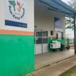 A imagem mostra a fachada de um prédio com uma pintura vibrante, dividida em verde na parte inferior e branco na superior, com uma faixa laranja separando as duas cores. À esquerda, um letreiro com o logo "Bem-Estar Animal" e o nome "Piracicaba" está fixado na parede. A entrada principal do prédio é de vidro, com portas duplas e um toldo cobrindo a área. À direita da entrada, um letreiro azul com as siglas "CCZ" e o nome "Piracicaba" está visível, assim como um extintor de incêndio vermelho. Uma área externa pavimentada com concreto, que apresenta algumas rachaduras e desníveis, leva à entrada. Ao fundo, uma cerca e vegetação compõem o cenário. O céu azul com algumas nuvens completa a cena.