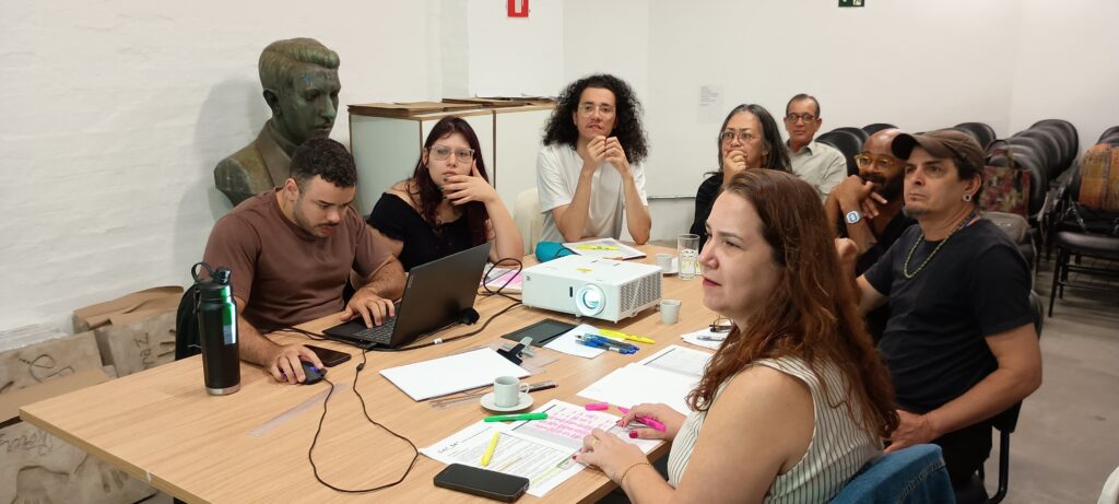 A imagem mostra um grupo de pessoas reunidas em torno de uma mesa de madeira em uma sala com paredes brancas. Há um projetor sobre a mesa, voltado para a frente, e diversos papéis espalhados. No primeiro plano, à esquerda, um homem com barba usa um laptop e um mouse. Ao seu lado, uma mulher com óculos escuros e cabelos cacheados está sentada, com a mão no queixo. Mais atrás, um homem com cabelos cacheados e óculos observa atentamente. No lado direito da mesa, uma mulher com cabelos longos e castanhos está sentada, olhando para a frente, com uma caneta rosa na mão. Outras pessoas estão sentadas ao redor da mesa, algumas em foco e outras mais desfocadas, indicando que a imagem foi tirada em um ambiente de reunião ou workshop. Ao fundo, há um busto de bronze e cadeiras empilhadas.