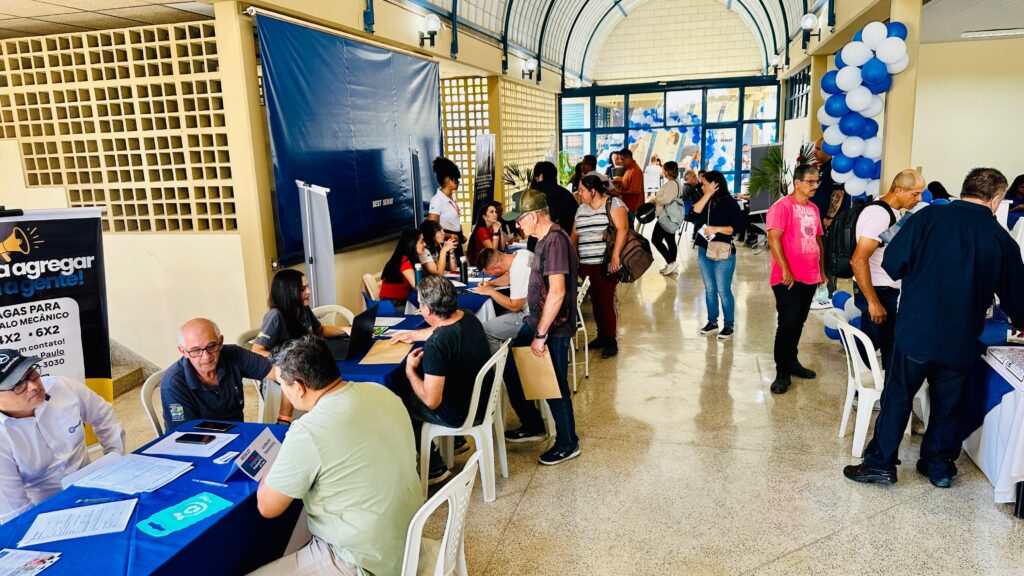 A imagem retrata um evento de recrutamento ou feira de empregos, com várias pessoas interagindo em um ambiente interno. Há mesas com voluntários ou recrutadores e candidatos conversando, preenchendo formulários ou olhando informações. Em destaque, uma mesa à esquerda com um banner que anuncia "Vagas para Mecânico", com detalhes de contato. Pessoas estão sentadas e em pé ao redor desta e de outras mesas. Ao fundo, há uma parede com um padrão quadriculado e um grande banner azul com o logo "SEST SENAT". Balões azuis e brancos decoram o espaço, conferindo um tom festivo ao evento. A iluminação é clara, vindo de cima, e o piso é de granilite.
