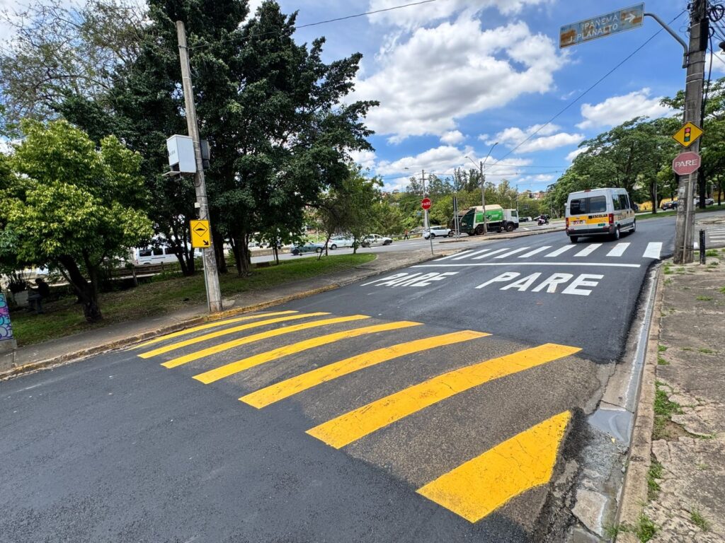 A imagem mostra uma rua com uma faixa de pedestres recém-pintada com listras amarelas proeminentes e a palavra "PARE" escrita em branco no asfalto. Há um redutor de velocidade (lombada) também marcado com listras amarelas. No lado direito da rua, há um poste com um semáforo e placas de trânsito, incluindo uma placa vermelha de "PARE" e uma placa amarela com o símbolo de um obstáculo na via. Um ônibus escolar branco com a palavra "ESCOLAR" escrita na lateral está parado próximo à faixa de pedestres. Ao fundo, vê-se uma área com árvores e vegetação, e mais adiante, outros veículos e edifícios. O céu está parcialmente nublado, com nuvens brancas e volumosas sobre um fundo azul. A cena parece ser de um dia claro e ensolarado.