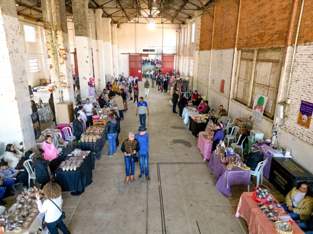 A imagem mostra uma visão aérea de um mercado ou feira de artesanato. Várias barracas estão dispostas em corredores, vendendo diversos produtos como roupas, joias, canecas e outros itens artesanais. Pessoas circulam entre as barracas, algumas comprando, outras conversando e outras trabalhando. O espaço parece ser um galpão amplo com colunas de cimento visíveis. A iluminação é natural, vinda de janelas altas e da entrada do galpão.