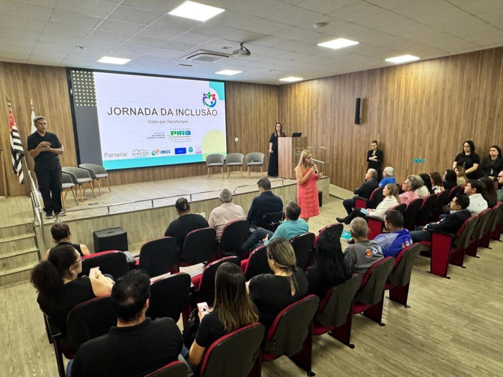 A imagem mostra um evento chamado "Jornada da Inclusão: Cotas que Transformam". Há um palestrante em pé à esquerda, em frente a uma tela grande que exibe o título do evento e logotipos. No palco, uma mulher de vestido rosa está falando em um microfone, enquanto outra mulher em um vestido preto está atrás de um púlpito. O público, composto por homens e mulheres de diversas idades, está sentado em cadeiras arranjadas em fileiras, assistindo à apresentação. O ambiente parece ser uma sala de palestras ou auditório, com paredes de madeira e iluminação adequada. Há bandeiras de São Paulo à esquerda do palco.