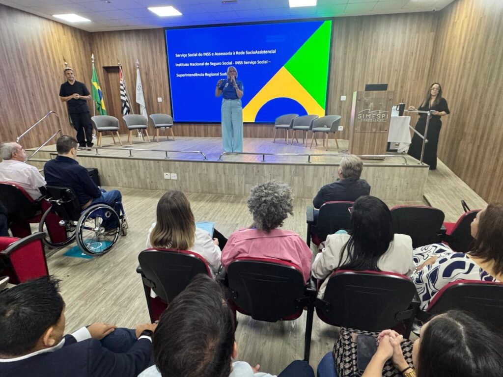 A imagem mostra uma sala de conferências com um palco elevado. No palco, há um homem de pé à esquerda, vestindo uma camisa preta e calças escuras, e uma mulher de pé à direita, usando um microfone e falando para a audiência. Atrás deles, uma grande tela exibe o texto "Serviço Social do INSS e Assessoria à Rede SocioAssistencial - Instituto Nacional do Seguro Social - INSS Serviço Social - Superintendência Regional Sud", com as cores da bandeira do Brasil ao fundo. Na plateia, sentada em cadeiras, há diversas pessoas. Notavelmente, um homem em uma cadeira de rodas está posicionado nas primeiras fileiras. As pessoas na plateia parecem atentas à apresentação. A sala possui um ambiente formal, com paredes de madeira e iluminação adequada para eventos. Há também um púlpito com o logotipo "SIMESPI" à direita do palco.