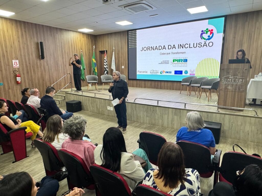 A imagem mostra uma sala de eventos com um palco e uma plateia sentada. No palco, há um telão exibindo a frase "JORNADA DA INCLUSÃO" e logos de patrocinadores. Dois homens estão em pé no palco, um deles falando ao microfone. Várias pessoas estão sentadas na plateia, prestando atenção à apresentação. As paredes da sala são revestidas de madeira, e há cadeiras vermelhas dispostas em fileiras. Bandeiras do Brasil e de São Paulo podem ser vistas no fundo do palco.