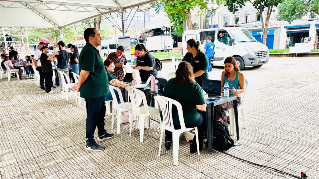 A imagem mostra um evento ao ar livre, possivelmente uma feira de saúde ou um posto de vacinação, sob uma tenda branca. Várias pessoas estão reunidas, algumas sentadas em cadeiras brancas de plástico e outras em pé. Há mesas com laptops e outros equipamentos, sugerindo que atividades estão sendo realizadas. Ao fundo, é possível ver vans e edifícios, indicando um ambiente urbano. Em primeiro plano, um homem de camisa verde escura e calça preta está em pé, de costas para a câmera, olhando para o lado. Ao seu lado, uma mulher com cabelo ruivo e tatuagens nos braços está sentada, também de costas. Mais adiante, há um grupo de pessoas interagindo em torno de mesas com laptops. Uma mulher de top azul e calça estampada está sentada em uma das mesas, olhando para o laptop à sua frente. Outras pessoas vestindo camisetas pretas parecem estar trabalhando ou auxiliando os presentes.