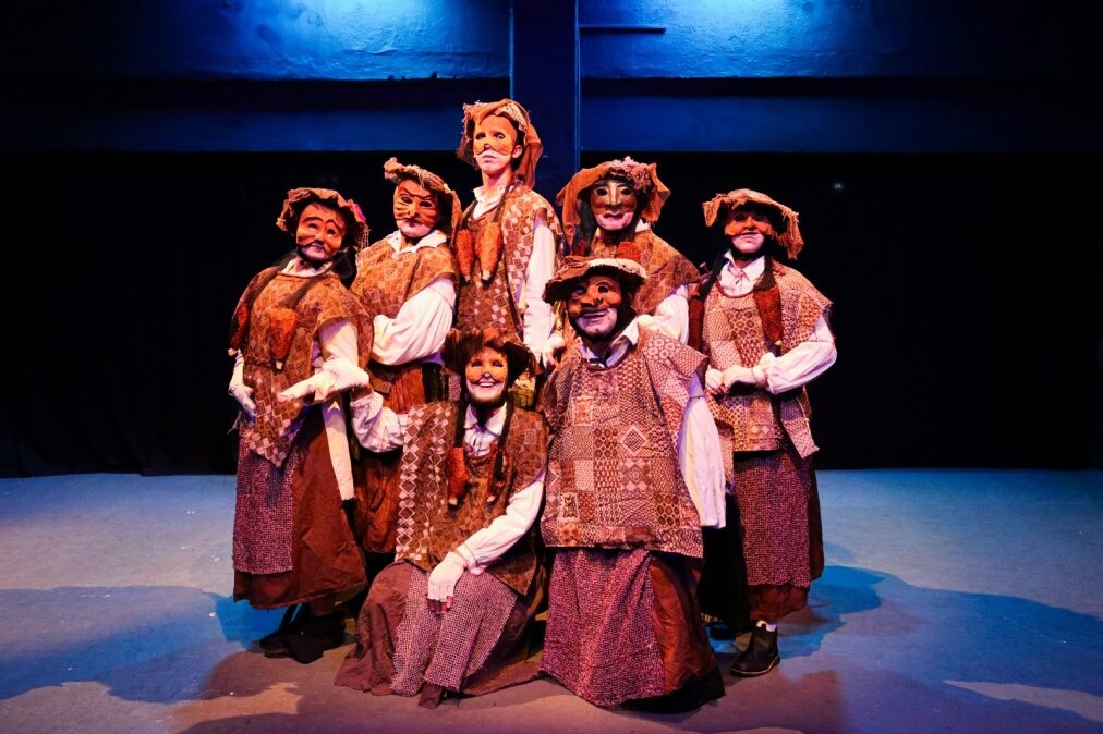 A imagem mostra um grupo de sete atores no palco, vestidos com trajes rústicos e usando máscaras. As máscaras cobrem seus rostos e são decoradas com padrões que lembram animais, como gatos ou raposas. Os trajes consistem em túnicas acolchoadas em tons de marrom e bege, com estampas geométricas e texturas variadas, sobrepostas a camisas brancas de mangas compridas e saias ou calças longas.A iluminação do palco é dramática, com um fundo escuro e um feixe de luz azul que ilumina a cena, criando sombras projetadas no chão. A composição do grupo sugere uma apresentação teatral, possivelmente uma peça folclórica ou infantil, dada a natureza lúdica das máscaras e dos figurinos. A disposição dos atores em diferentes níveis, alguns mais à frente e outros atrás, confere profundidade à imagem.