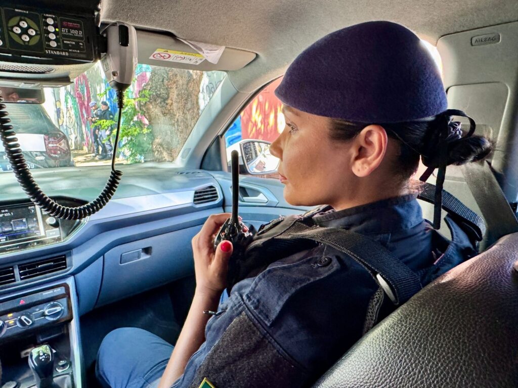 A imagem mostra uma mulher sentada no banco do passageiro de um carro, vestindo um uniforme azul escuro e um boina roxa. Ela está segurando um rádio comunicador preto na mão direita, com as unhas pintadas de vermelho. O interior do carro é visível, com o painel em tons de cinza e um rádio de carro com tela digital. No teto do carro, há um microfone de rádio preso com um fio espiralado. Através do para-brisa, é possível ver o exterior, onde há grafites coloridos em uma parede e duas pessoas vestindo uniformes semelhantes ao da mulher na imagem. Uma árvore com folhagem verde também é visível. A luz do sol ilumina a cena, criando sombras e realçando os detalhes.