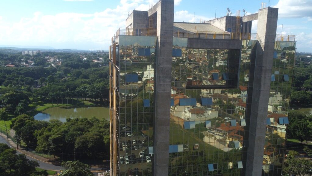 A imagem mostra uma vista aérea de um edifício moderno com fachada de vidro e estruturas de concreto aparentes. A fachada é composta por vários painéis de vidro que refletem o ambiente ao redor, criando um efeito visual interessante. Algumas janelas estão abertas, permitindo ver o interior ou o exterior do prédio. A estrutura de concreto tem um acabamento rústico, contrastando com o vidro liso e brilhante. Ao redor do edifício, há uma área verde bem cuidada, com gramados, árvores variadas, incluindo algumas com folhagem parecida com palmeiras, e um lago ou lagoa que adiciona um elemento natural ao cenário. Ao fundo, é possível ver uma cidade ou bairro com várias construções, sugerindo que o prédio está localizado em uma região urbana ou próxima a ela. O céu está claro, com poucas nuvens brancas, indicando um dia ensolarado. A perspectiva da foto é de um ponto alto, oferecendo uma visão ampla do edifício e do seu entorno, destacando a integração entre a arquitetura moderna e a natureza ao redor.