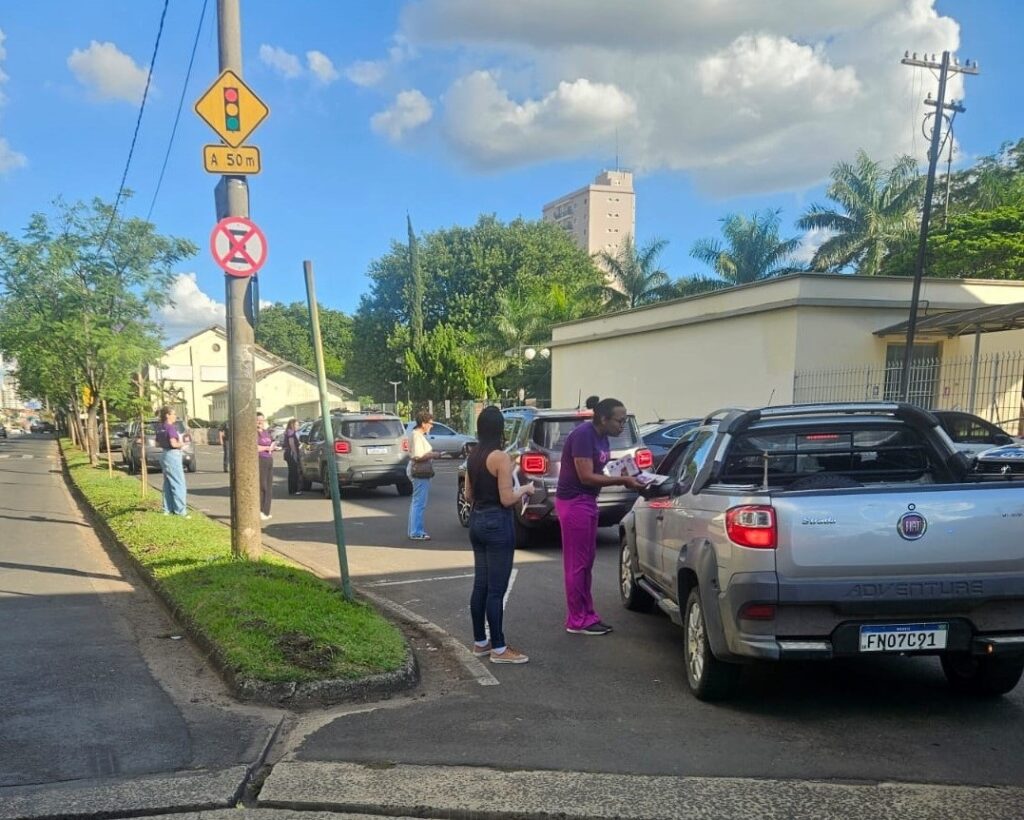 A imagem mostra uma rua com carros parados em fila, possivelmente em um semáforo ou em uma área de trânsito lento. Há várias pessoas em pé na calçada e próximas aos carros, algumas vestindo roupas roxas. Uma pessoa em pé perto de um carro prata está entregando algo para o motorista, que parece ser um panfleto ou folheto. Há também um poste com placas de trânsito, incluindo uma que indica um semáforo a 50 metros e outra que parece proibir estacionamento ou parada. O céu está azul com nuvens brancas, e há árvores verdes ao redor, sugerindo um dia ensolarado em um ambiente urbano.